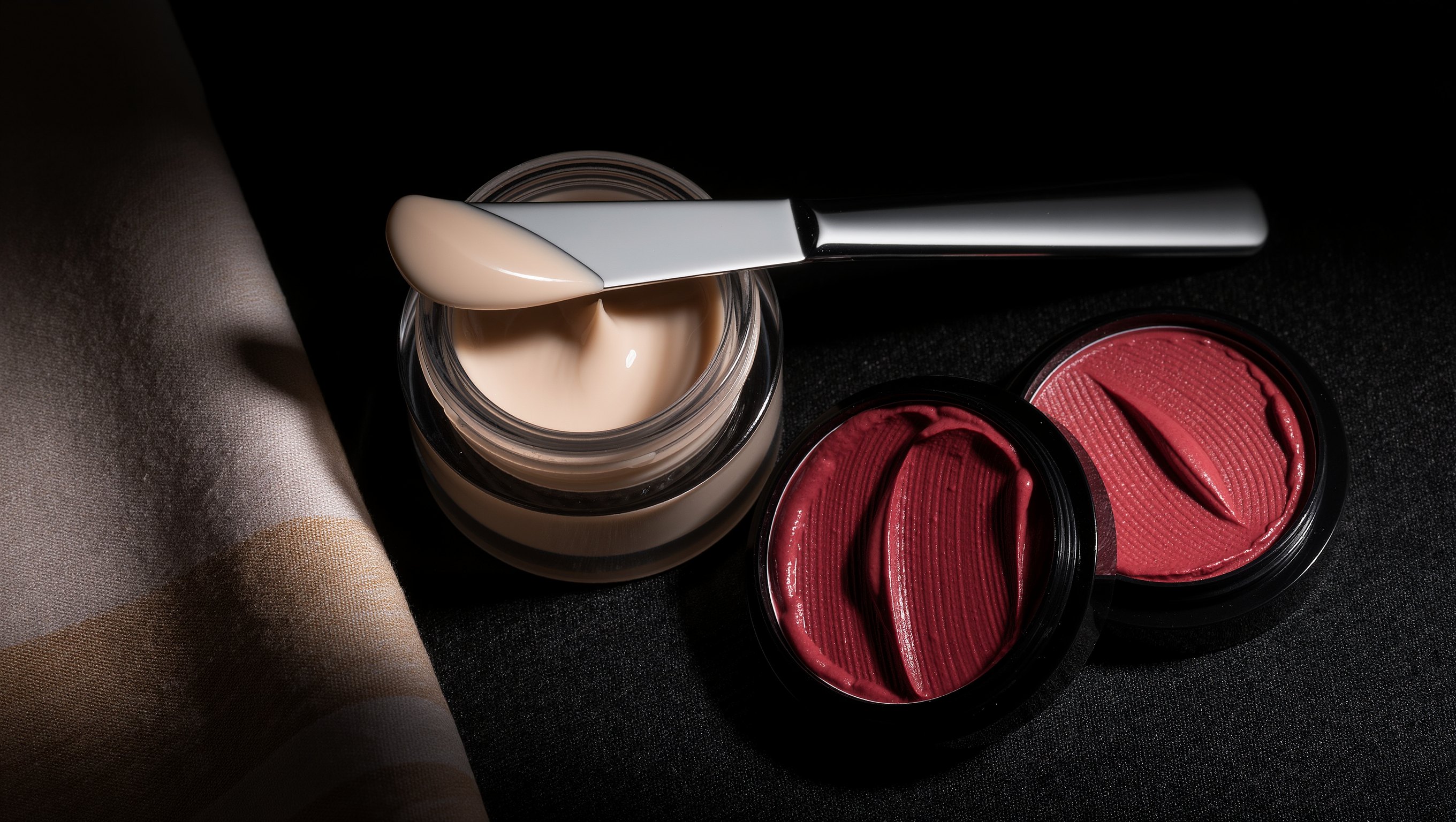 Cremerouge in einer offenen Dose neben einem Lippenstift in gedecktem Rot auf grobem Lodenstoff